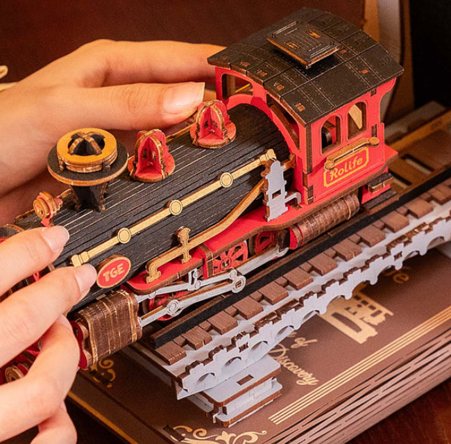 Hand holding a red and black toy train model on a wooden base with 'TGS' branding.