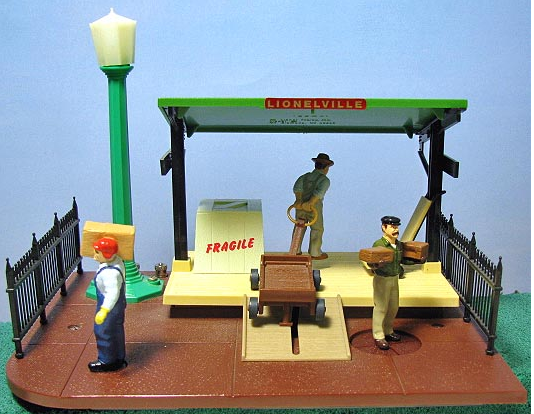 A toy model of an animated freight station with a green newsstand, a brown baggage handler figure, and a black lamp post with a light on top.