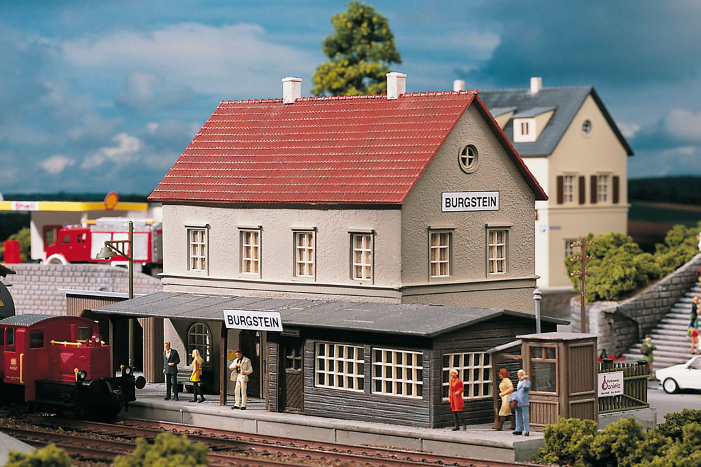HO-scale model of a Burgstein station building with a red roof, gray walls, and a platform. Model figures are visible on the platform, adding a human element to the scene.