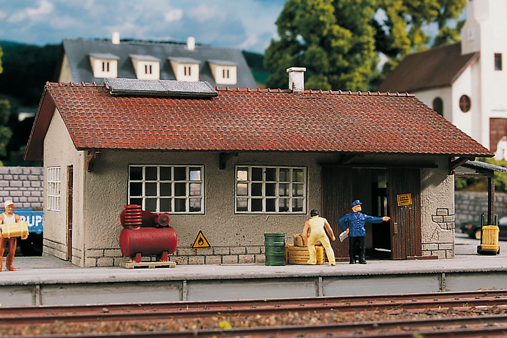 HO-scale model of a Burgstein goods depot with a red roof, gray walls, and a small model train engine in front.