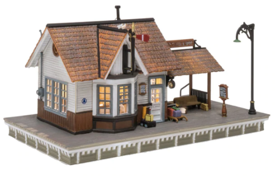 A model of a classic whistle stop depot with clapboard siding, a weathered cedar shake roof, a covered passenger waiting area, and an LED light. It includes a ticket clerk figure, luggage carts, benches, and a semaphore.