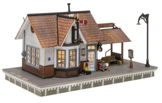 A model of a classic whistle stop depot with clapboard siding, a weathered cedar shake roof, a covered passenger waiting area, and an LED light. It includes a ticket clerk figure, luggage carts, benches, and a semaphore.