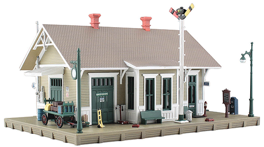 An N scale model of a two-story depot with detailed door and window styling, set on a platform with a train signal, luggage cart, and street lamps.