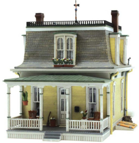 A model of a two-story Victorian-style house with a yellow front and white trim, featuring detailed architectural elements such as a weather vane, flowered window boxes, bracketed eaves, and a woodpile.