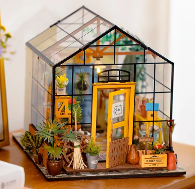 Miniature greenhouse model with plants and decorative items on a wooden surface.