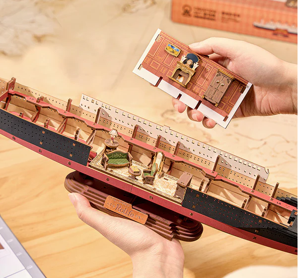 Model ship with detailed interior and hands holding a cutaway panel on a wooden surface