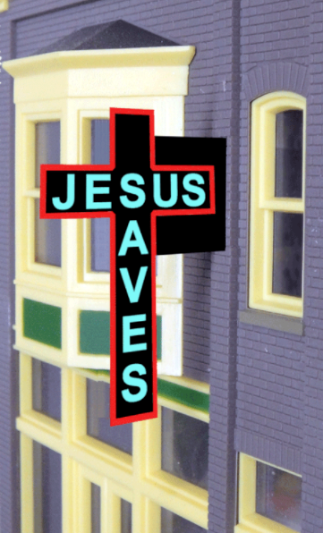 A novelty sign with a cross and the phrase 'JESUS SAVES' in capital letters on a red and black background, designed for use in a model train setup.