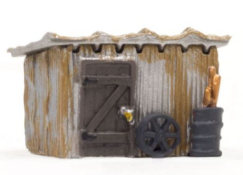 A model of a tin shack with rusted metal walls, a weather beaten door, and a corrugated metal roof. There is a barrel of iron and an old wheel leaning against the wall.