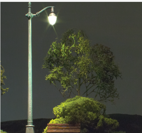 An arched cast iron street light with a warm white LED glow, placed next to a model of a tree and a bench, set against a dark background.