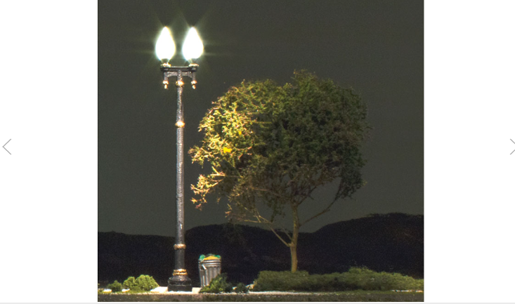 A HO Scale model train track with a double lamp post street light illuminated in warm white, placed next to a tree and shrubbery.